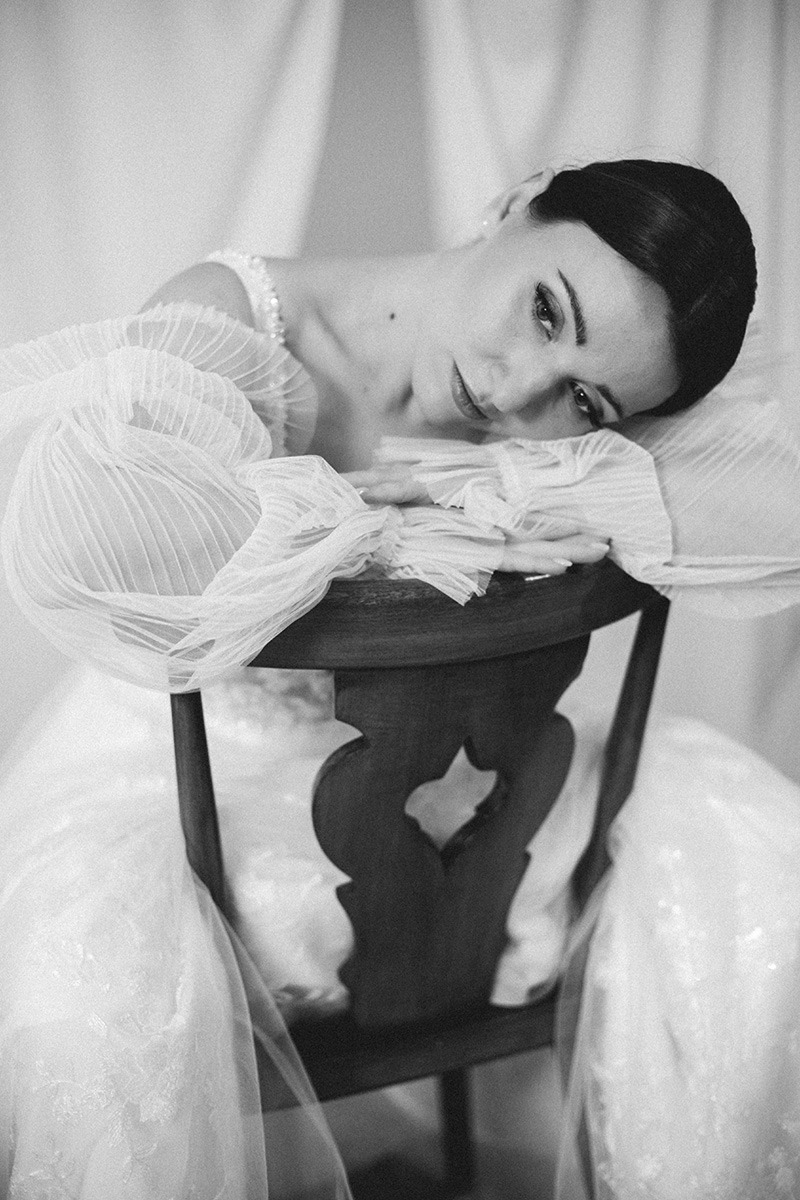 Braut mit professionellem Braut-Make-up und hochgesteckter Frisur, sitzend auf elegantem Stuhl – Styled Shooting für Hochzeit